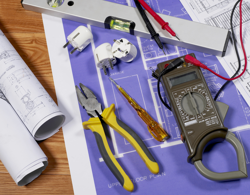 Electrical tools, blueprints, a voltage tester, and a clamp meter are arranged on a table, indicating work or planning for an electrical installation or repair.