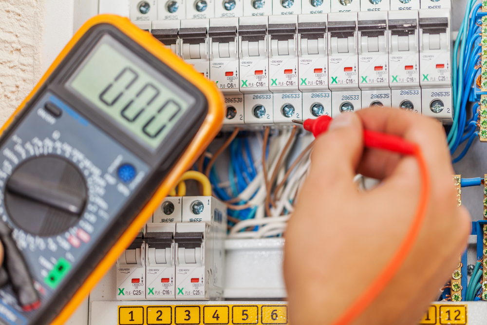 A person uses a digital multimeter to test electrical circuits inside a breaker panel with multiple wires and switches.