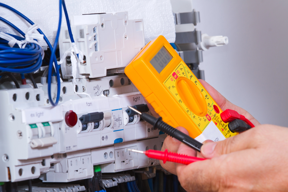 A person uses a digital multimeter to measure voltage on an electrical panel with various wires, switches, and circuit breakers.
