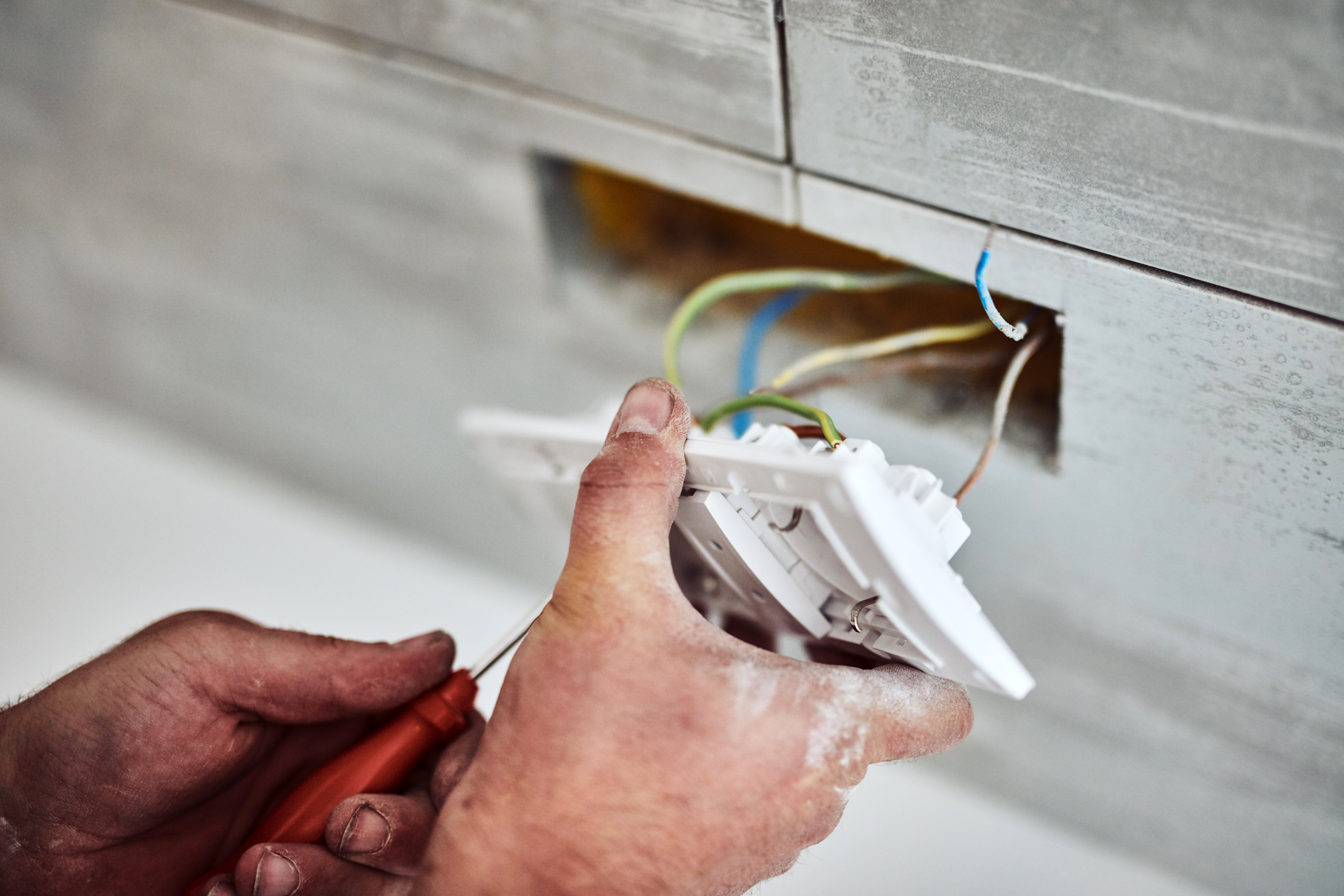 Hands installing or repairing a wall-mounted electrical outlet with exposed wires and a screwdriver against a tiled background.